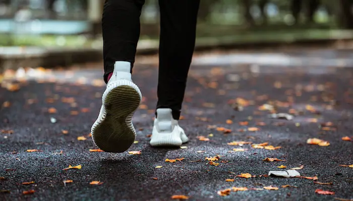 Feet of a person in white rubber shoes walking or jogging away on asphalt with orange leaves